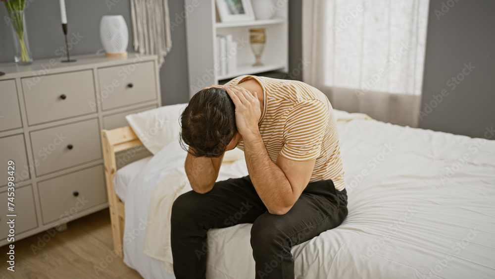 Upset young hispanic man sitting on bed in bedroom, holding his head in distress. Upset young hispanic man sitting on bed in bedroom, holding his head in distress.