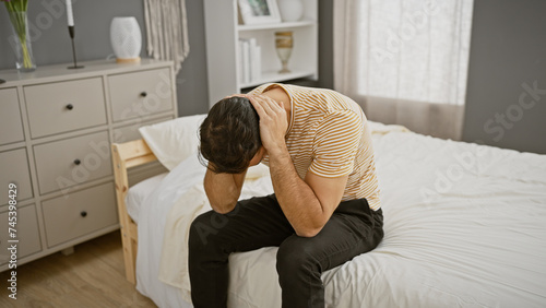 Upset young hispanic man sitting on bed in bedroom, holding his head in distress.