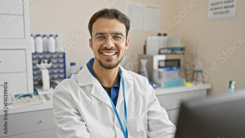 Handsome young hispanic man with beard smiling in laboratory wearing lab coat and glasses