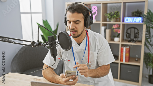 Handsome hispanic man in medical uniform podcasting with microphone and smartphone in radio studio. photo