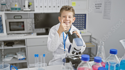 Adorable little blond boy  a budding scientist  giving a thumb up gesture while confidently using a microscope in a medical lab  all smiles amid his exciting scientific discovery.