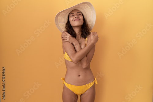Young hispanic woman wearing bikini and summer hat hugging oneself happy and positive  smiling confident. self love and self care