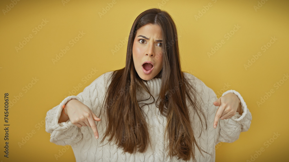 Surprised young hispanic woman with open mouth pointing downwards, wearing a sweater against a yellow background. Surprised young hispanic woman with open mouth pointing downwards, wearing a sweater against a yellow background.