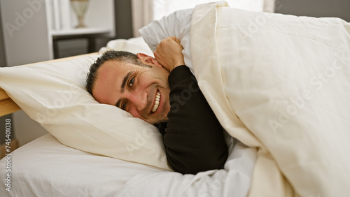 A smiling young man nestled in bed sheets expressing comfort and relaxation in a cozy bedroom setting. © Krakenimages.com