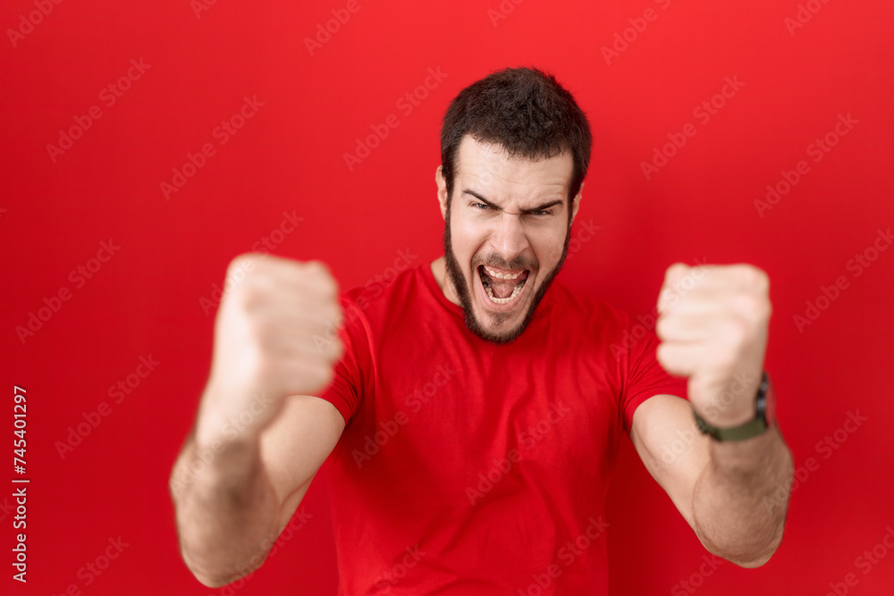 Young hispanic man wearing casual red t shirt angry and mad raising fists frustrated and furious while shouting with anger. rage and aggressive concept. Young hispanic man wearing casual red t shirt angry and mad raising fists frustrated and furious while shouting with anger. rage and aggressive concept.