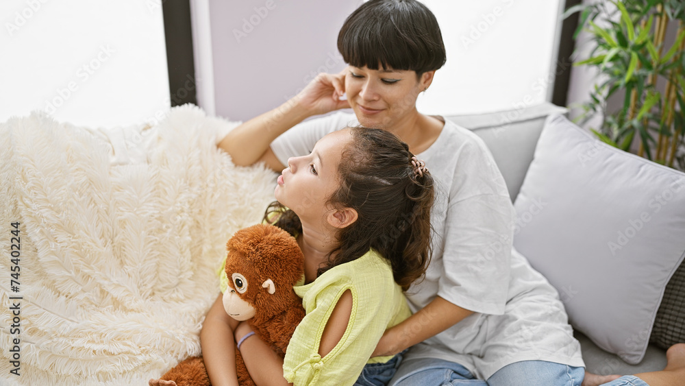 Lovely mother and daughter share a joyful moment hugging a funny monkey doll while sitting casually on the living room sofa at home, expressing the positive and confident happiness of family love. Lovely mother and daughter share a joyful moment hugging a funny monkey doll while sitting casually on the living room sofa at home, expressing the positive and confident happiness of family love.