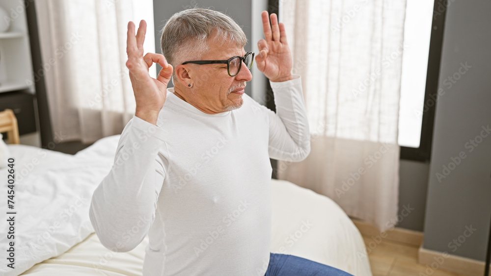Fototapeta premium A middle-aged man with grey hair, wearing glasses, makes gestures sitting on a bed, indoors, portraying a relaxed or playful mood.