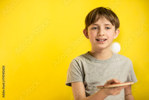 portrait of a smiling 11 year old boy, with a racket and a table tennis ball, on a yellow background.