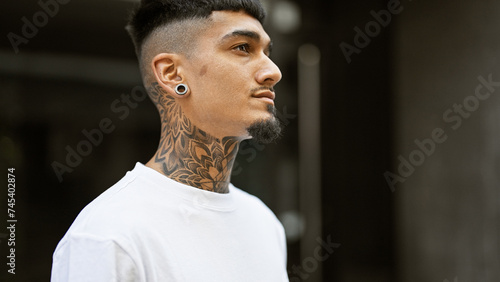 Cool, tattooed young latin man giving a serious look, outdoors. a relaxed pose on a sunny city street, concentrating on his urban lifestyle.