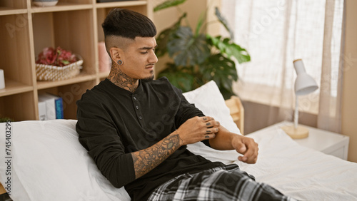 Photos Handsome young latin man, sitting on his bedroom bed, feeling the itch of dermatitis while scratching his itchy tattooed arm