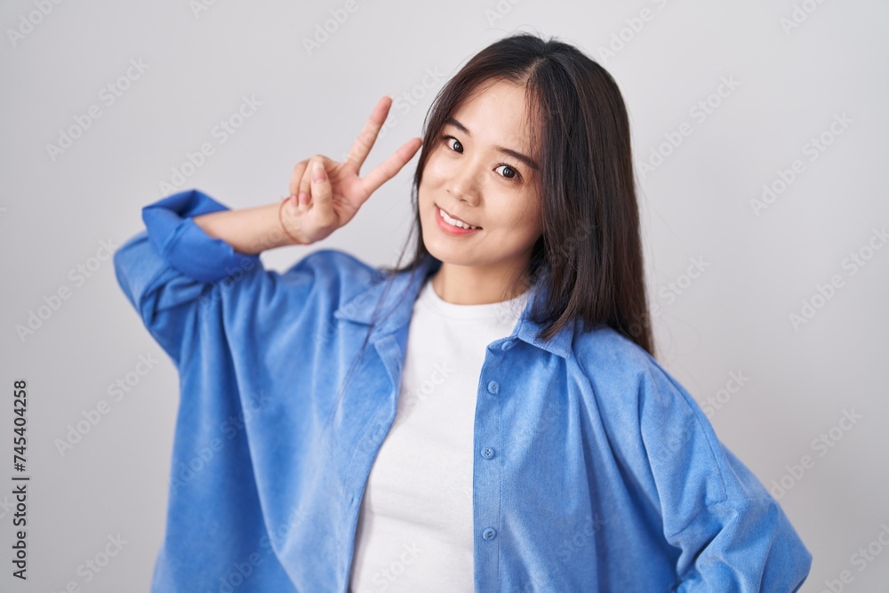 Young chinese woman standing over white background smiling looking to the camera showing fingers doing victory sign. number two. Young chinese woman standing over white background smiling looking to the camera showing fingers doing victory sign. number two.