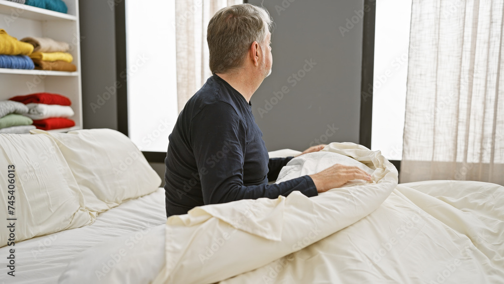 A grey-haired man sits contemplatively on a neatly made bed in a modern bedroom with a wardrobe and curtains in the background. A grey-haired man sits contemplatively on a neatly made bed in a modern bedroom with a wardrobe and curtains in the background.