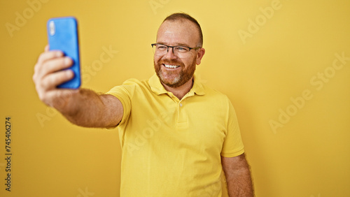 Confident, smiling caucasian man standing casual, making a fun selfie with smartphone against isolated yellow background. his cool glasses and attractive beard add confidence to his happy expression.