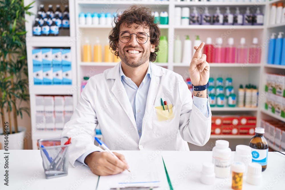 Hispanic young man working at pharmacy drugstore gesturing finger crossed smiling with hope and eyes closed. luck and superstitious concept.