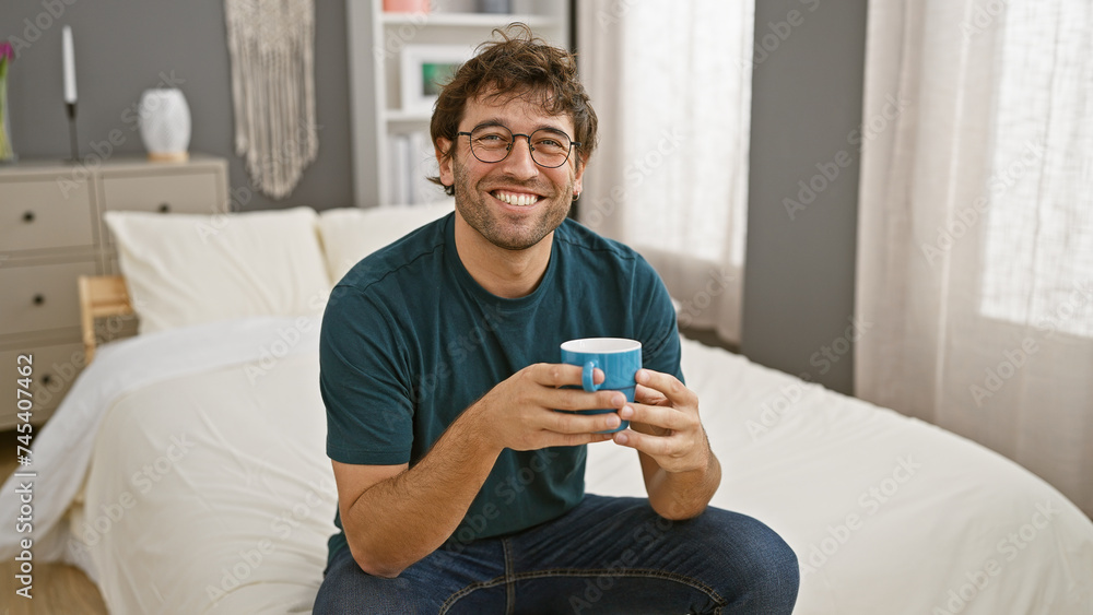 Smiling young hispanic man with beard holding a mug in a cozy bedroom setting. Smiling young hispanic man with beard holding a mug in a cozy bedroom setting.