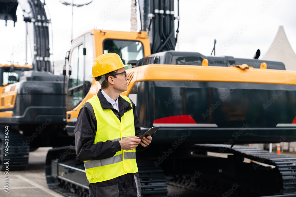 Engineer in a helmet with a digital tablet stands next to construction excavators.. Engineer in a helmet with a digital tablet stands next to construction excavators..