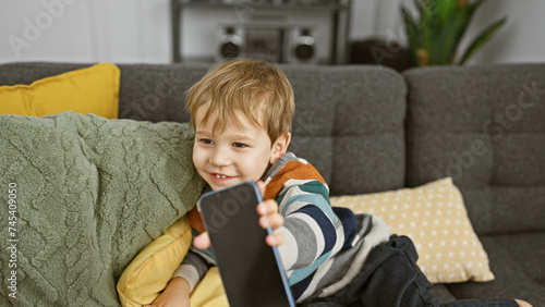 A cheerful blond toddler boy holding a smartphone while sitting on a couch with pillows in a cozy living room.