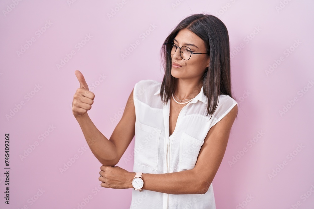 Brunette young woman standing over pink background wearing glasses looking proud, smiling doing thumbs up gesture to the side Brunette young woman standing over pink background wearing glasses looking proud, smiling doing thumbs up gesture to the side