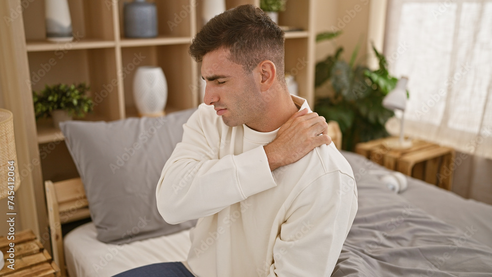 Young hispanic man suffering for shoulder injury sitting on bed at bedroom