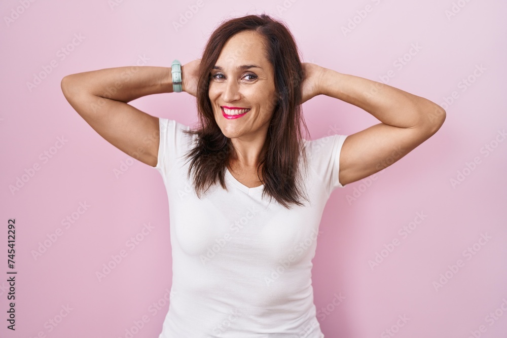 Middle age brunette woman standing over pink background relaxing and stretching, arms and hands behind head and neck smiling happy Middle age brunette woman standing over pink background relaxing and stretching, arms and hands behind head and neck smiling happy