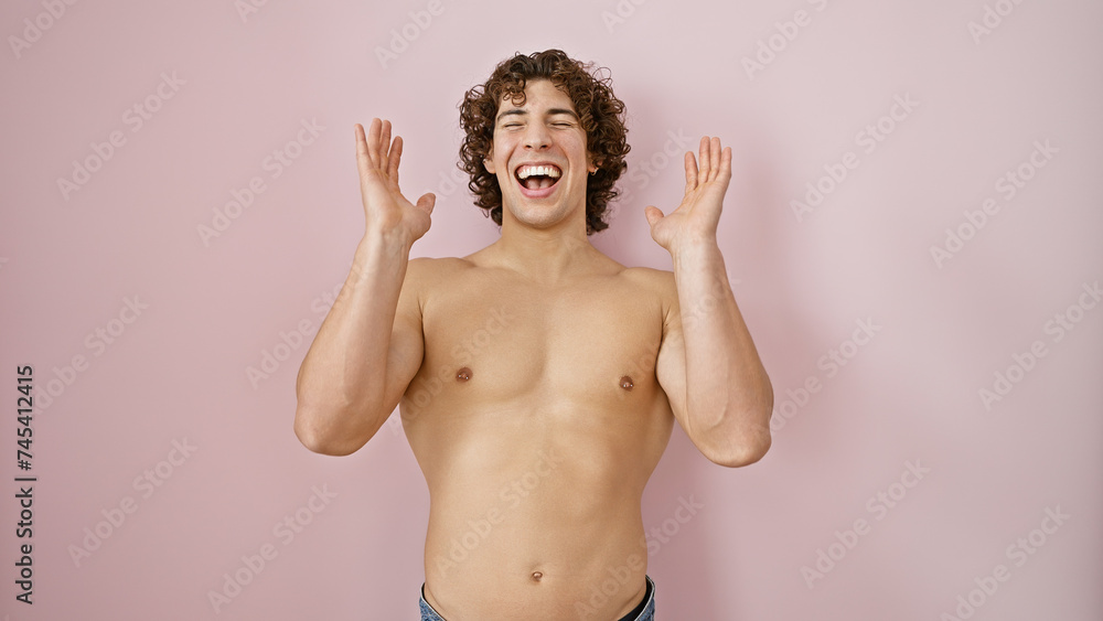 Fototapeta premium A joyful young man with curly hair rejoices shirtless against a pink background, embodying energy and vivacity.
