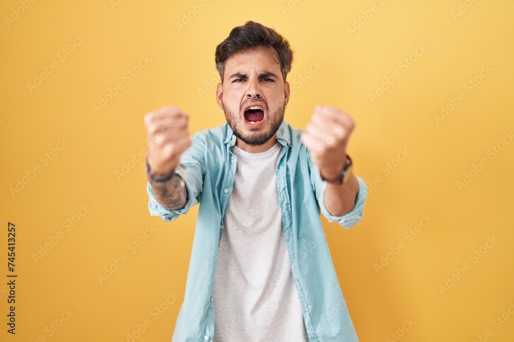 Young hispanic man with tattoos standing over yellow background angry and mad raising fists frustrated and furious while shouting with anger. rage and aggressive concept. Young hispanic man with tattoos standing over yellow background angry and mad raising fists frustrated and furious while shouting with anger. rage and aggressive concept.