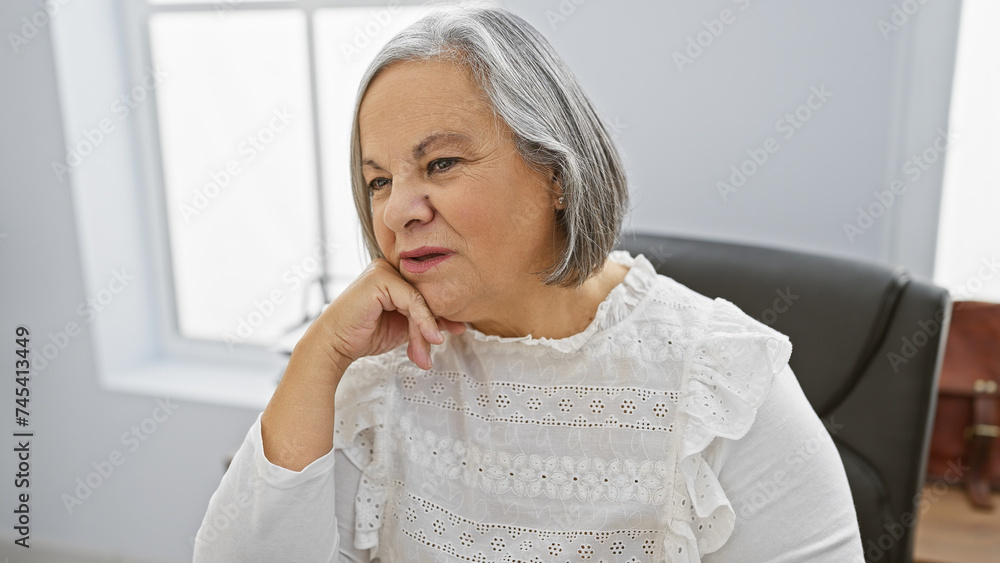 A contemplative senior woman with grey hair, wearing a white blouse, seated indoors. A contemplative senior woman with grey hair, wearing a white blouse, seated indoors.