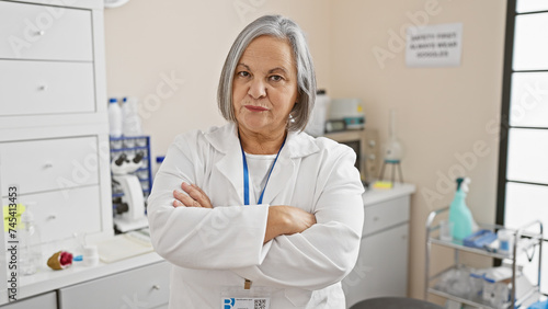 Confident senior woman scientist with grey hair stands with arms crossed in a laboratory setting.
