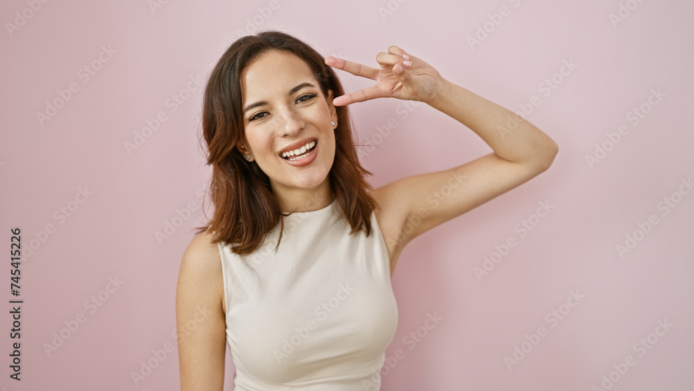 A cheerful young hispanic woman makes a peace sign against an isolated pink background, exuding confidence and beauty. A cheerful young hispanic woman makes a peace sign against an isolated pink background, exuding confidence and beauty.