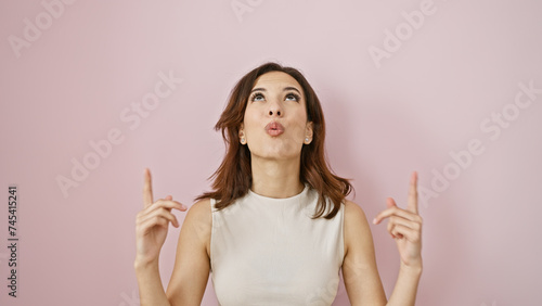 Young hispanic woman with a playful expression pointing upwards against a pink wall, evoking a light-hearted vibe.