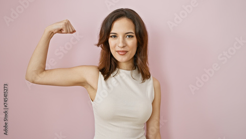 Confident young hispanic woman flexing arm muscles against a soft pink background, portraying strength and femininity.
