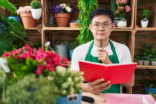 Young chinese man florist reading notebook with doubt expression at flower shop