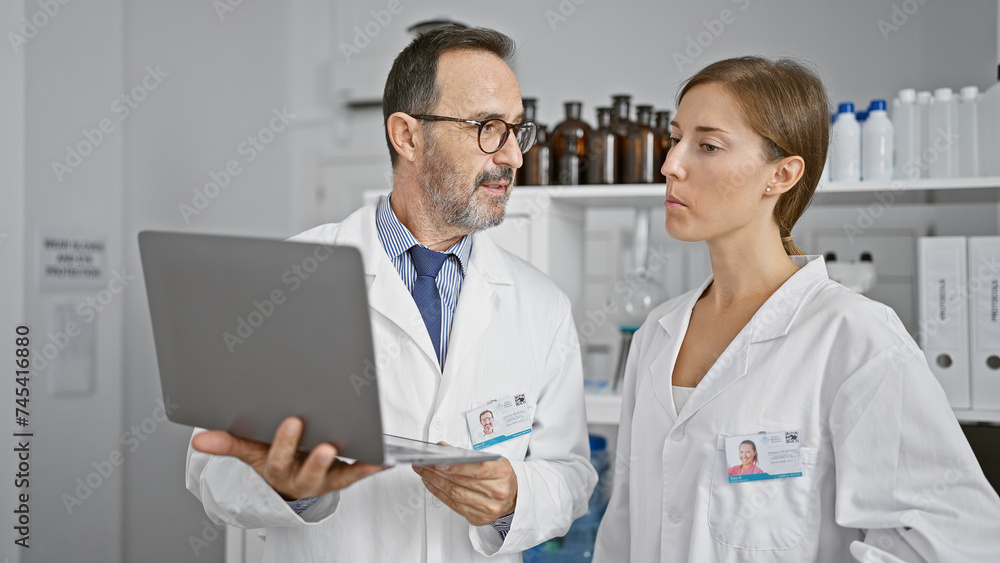 Fototapeta premium Two dedicated scientists working shoulder-to-shoulder, speaking over lab analysis on a laptop in the heart of a bustling medical research laboratory.