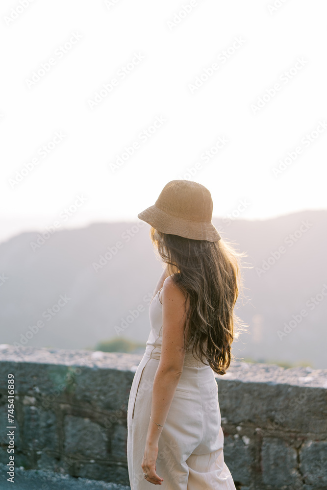 Young woman in a hat stands half-turned near a stone fence and looks at the mountains