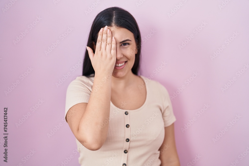 Young hispanic woman standing over pink background covering one eye with hand, confident smile on face and surprise emotion. Young hispanic woman standing over pink background covering one eye with hand, confident smile on face and surprise emotion.