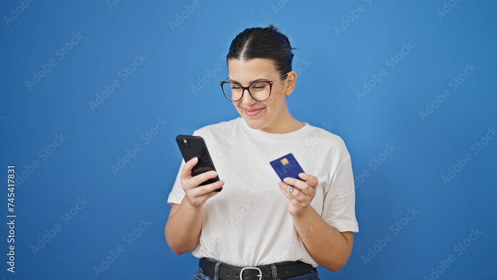 Young beautiful hispanic woman using smartphone and credit card over isolated blue background Young beautiful hispanic woman using smartphone and credit card over isolated blue background