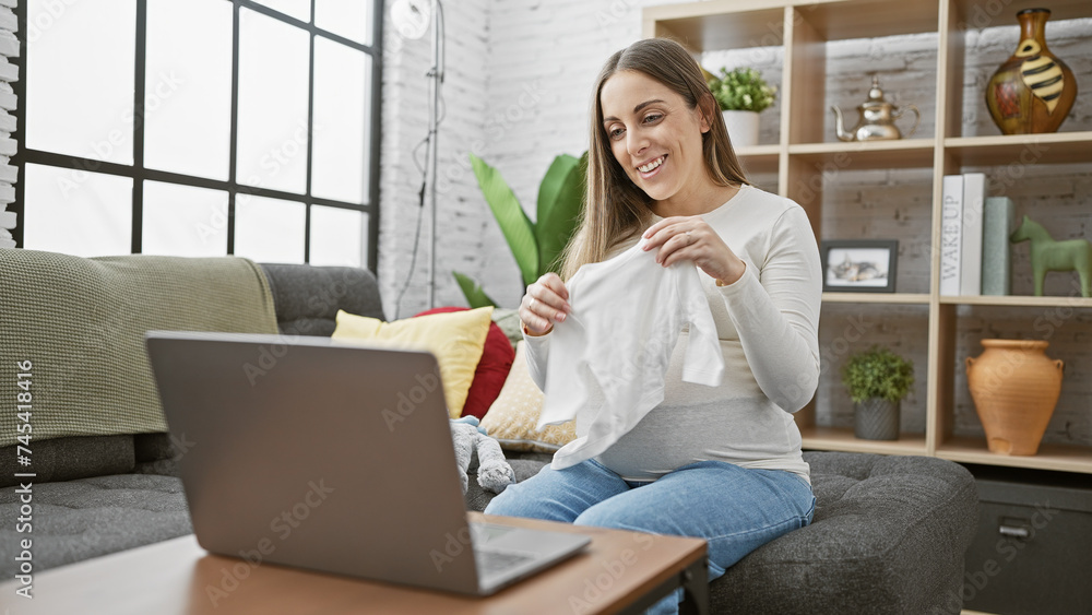 Smiling pregnant woman holding baby clothes during a video call at home. Smiling pregnant woman holding baby clothes during a video call at home.