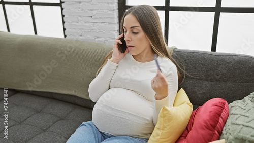 A pregnant hispanic woman sitting indoor on a couch, talking on the phone with a concerned expression