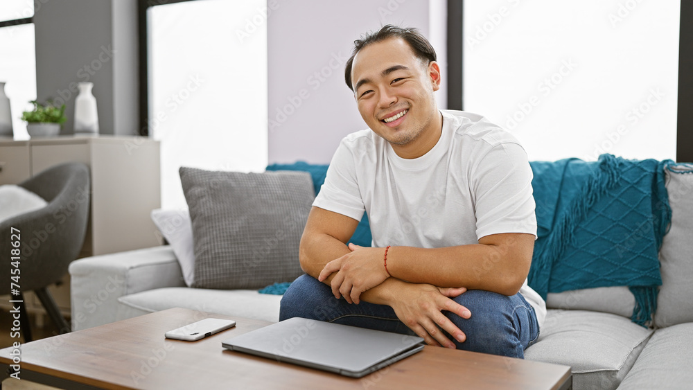 Cheerful young chinese man, devoted to his laptop, reveling in the comfort of his sofa at home in classic pigtail hairstyle Cheerful young chinese man, devoted to his laptop, reveling in the comfort of his sofa at home in classic pigtail hairstyle