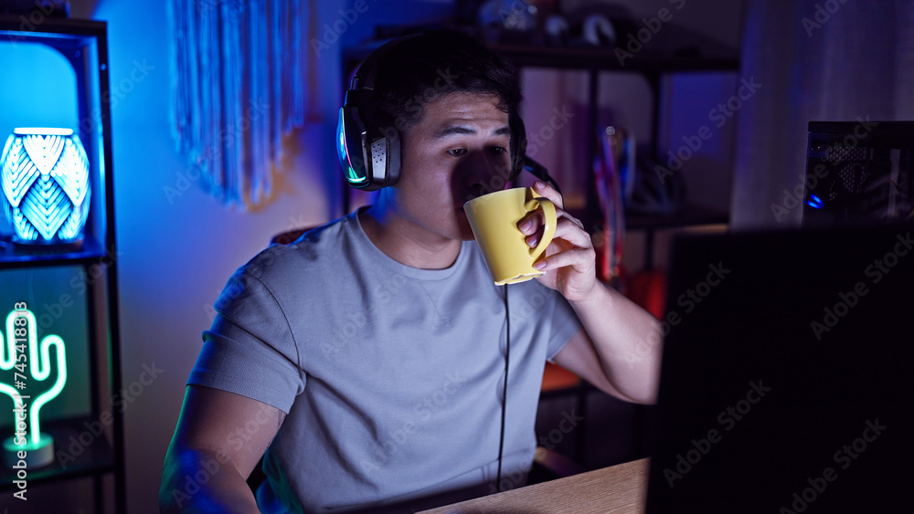 A young asian man enjoys a beverage in a cozy gaming room at night, illuminated by colorful neon lights.