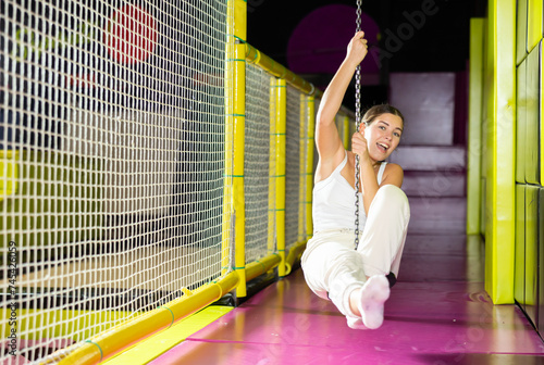 Young girl in white sportswear having fun in modern indoor trampoline amusement center, riding zip line against vibrant colorful background..