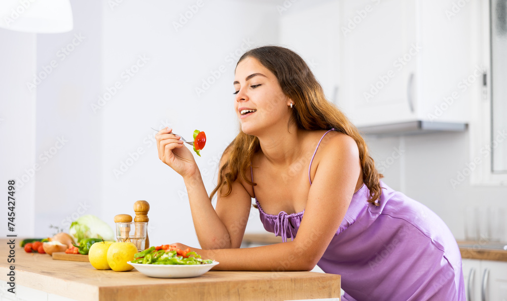 Portrait of positive girl in purple nightgown tasting vegetable salad at kitchen