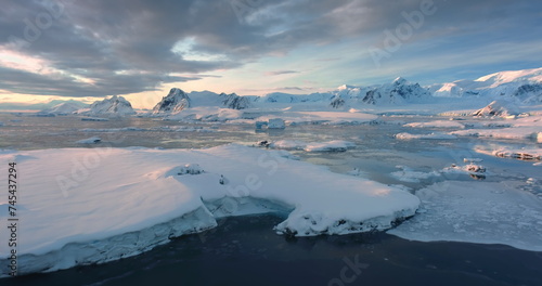 Fly above frozen panorama of an Antarctica. Snow covered mountain range  ice ocean water  icebergs  glaciers  sunset clouds sky. Beauty of wild untouched nature. Travel South Pole. Aerial drone shot