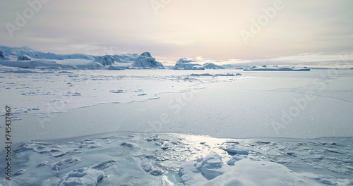 Fly over frozen Antarctic ocean sunset landscape. Snow covered untouched wilderness of South Pole. Desert white land of snow and ice aerial drone shot. Mountains in background. Nature conservation