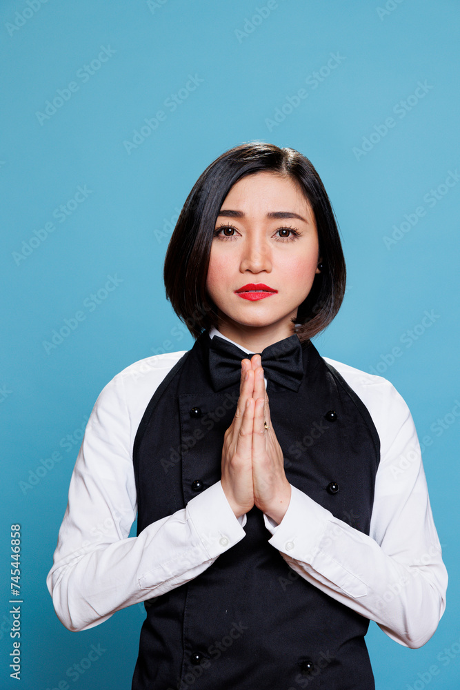 Young asian woman receptionist meditating with folded palms, relaxing and looking at camera. Attractive cafe waitress wearing uniform standing in zen pose portrait on blue background Young asian woman receptionist meditating with folded palms, relaxing and looking at camera. Attractive cafe waitress wearing uniform standing in zen pose portrait on blue background