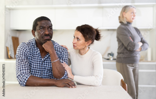 Middle-aged woman trying to calm angry man sitting at the kitchen table while mature woman quarreling to him standing behind