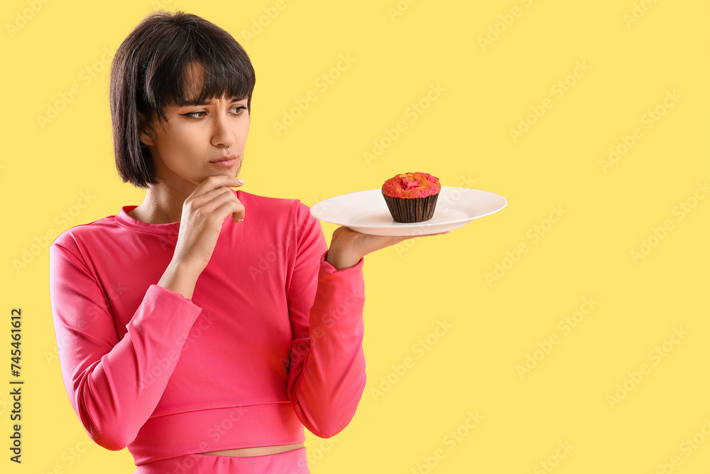Thoughtful sporty young woman with muffin on yellow background. Diet concept Thoughtful sporty young woman with muffin on yellow background. Diet concept