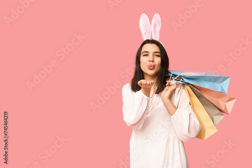 Beautiful young woman in bunny ears with shopping bags blowing kiss on pink background. Easter celebration