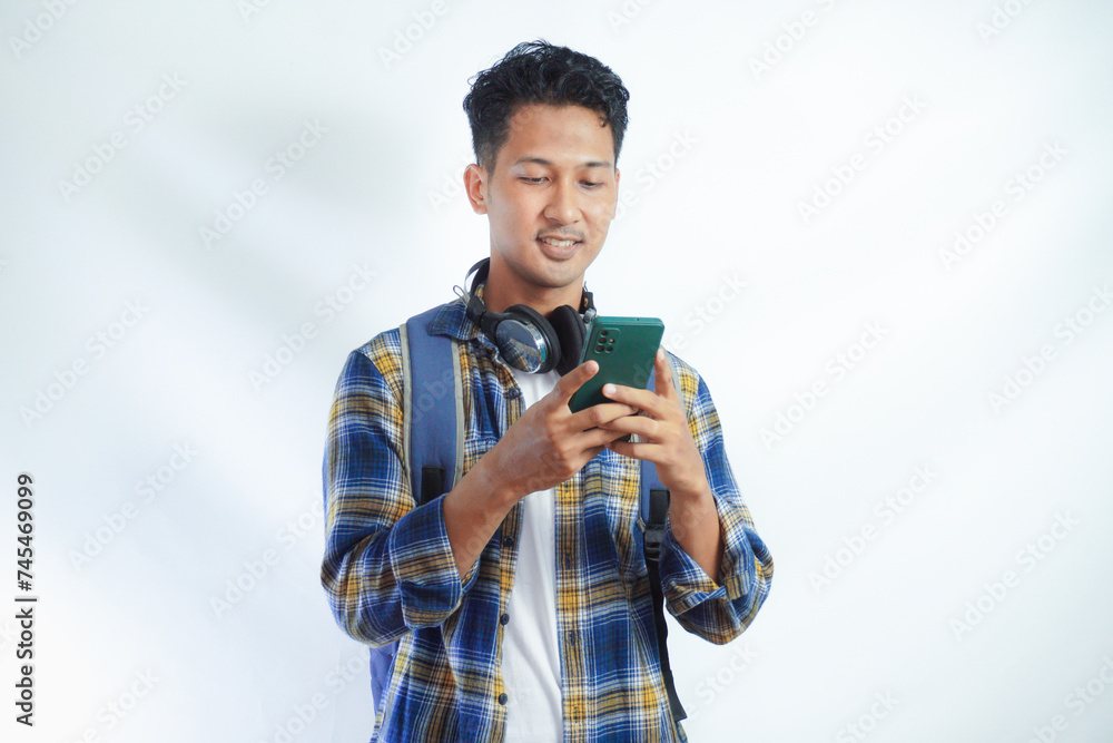 Smiling happy young Asian man student in casual clothes backpack using mobile phone, chatting online and reading news isolated on yellow background. high school university college concept Smiling happy young Asian man student in casual clothes backpack using mobile phone, chatting online and reading news isolated on yellow background. high school university college concept
