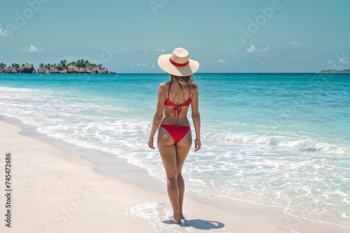 Back view of young woman in swimsuit and straw hat on tropical beach. A woman in a bikini stands on a beach.